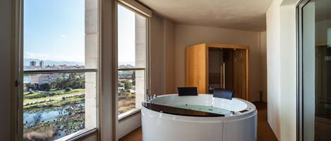 A modern bathroom with a freestanding bathtub, large windows offering a view of a cityscape and greenery, and a wooden floor.