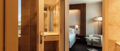 A hotel room with a glass door leading to a bathroom, a bed with white linens, a bedside table with a lamp, and a small white table.