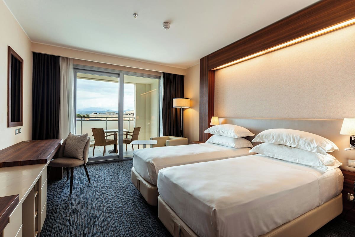 A hotel room with two beds, a desk, and a chair. There is a view of the city through the window.