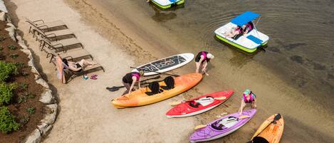 Una playa con kayaks y tablas de remo, una fila de sillas de descanso y una lancha con toldo.