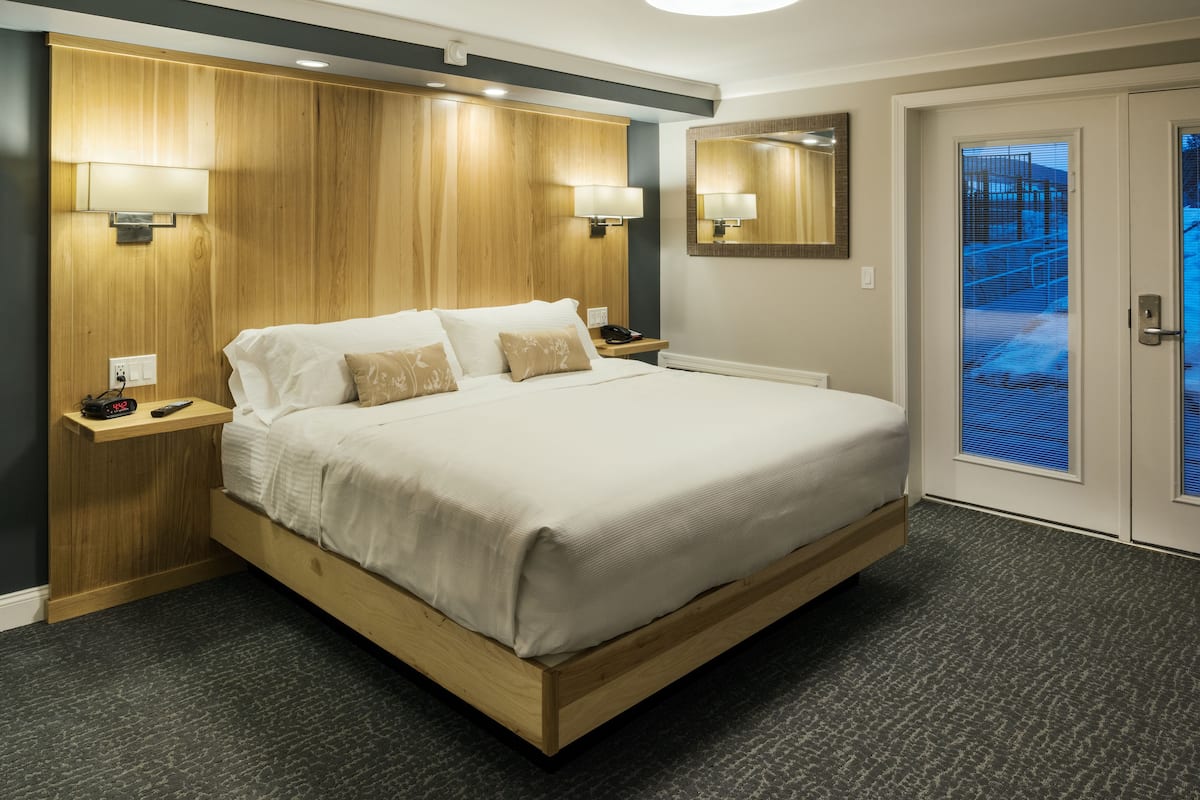 A hotel room with a large bed, wooden headboard, two bedside tables with lamps, a mirror, and a door leading to a balcony.