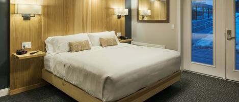 A hotel room with a large bed, wooden headboard, two bedside tables with lamps, a mirror, and a door leading to a balcony.