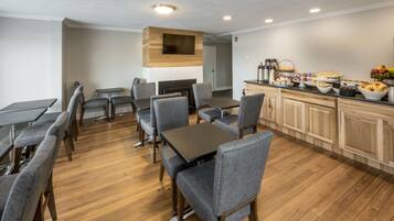 A modern dining area with wooden tables and grey upholstered chairs, a fireplace, and a breakfast bar with various food items.
