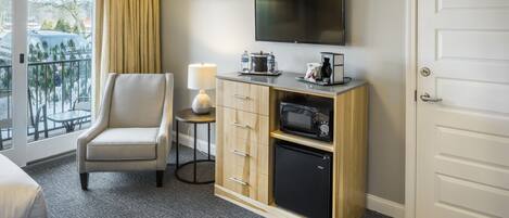 Habitación de hotel con cama, una silla, televisor, microondas, refrigerador, una mesita con lámpara y una puerta.