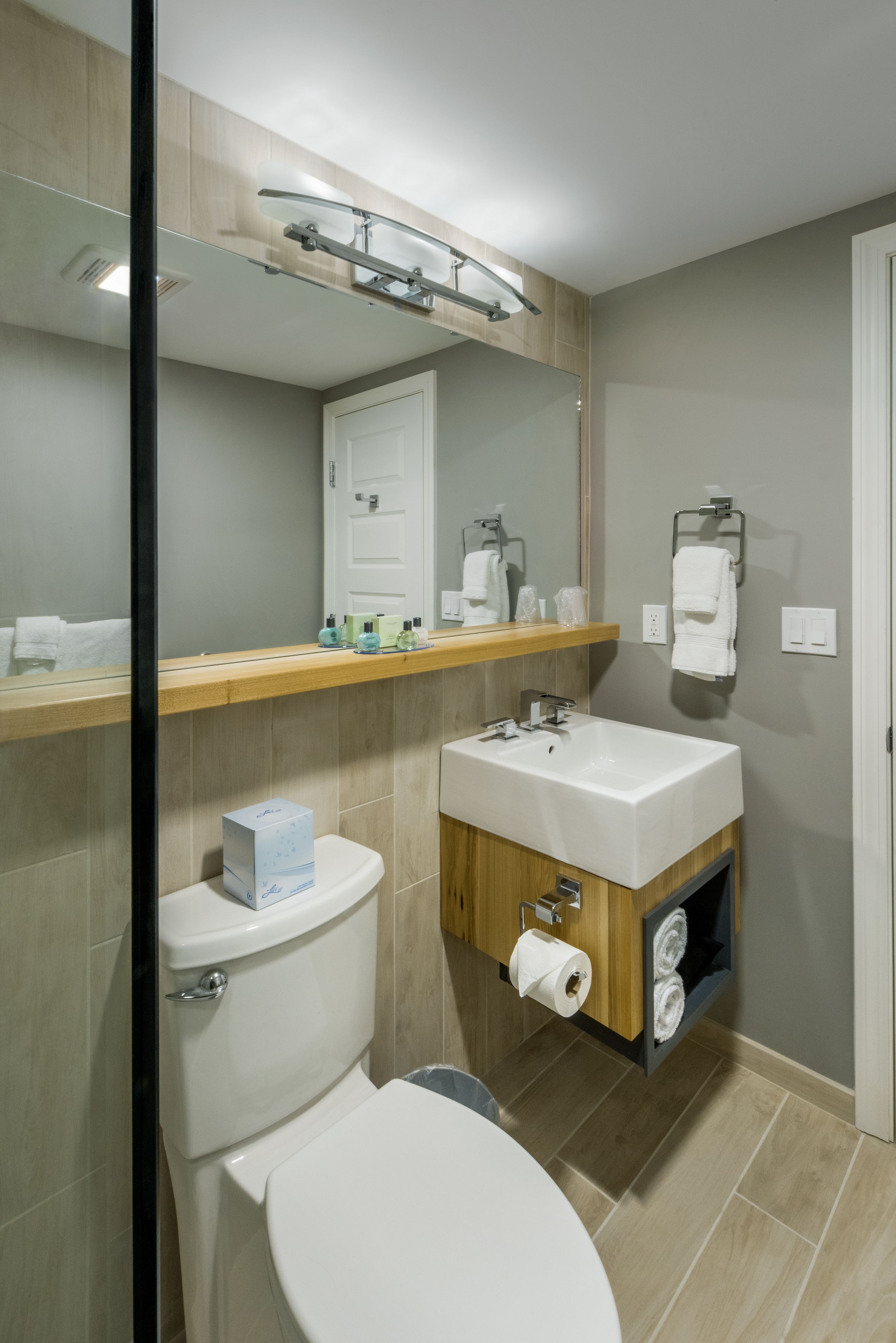 A modern bathroom with a toilet, sink, and mirror. There is a towel rack and a shelf with toiletries.