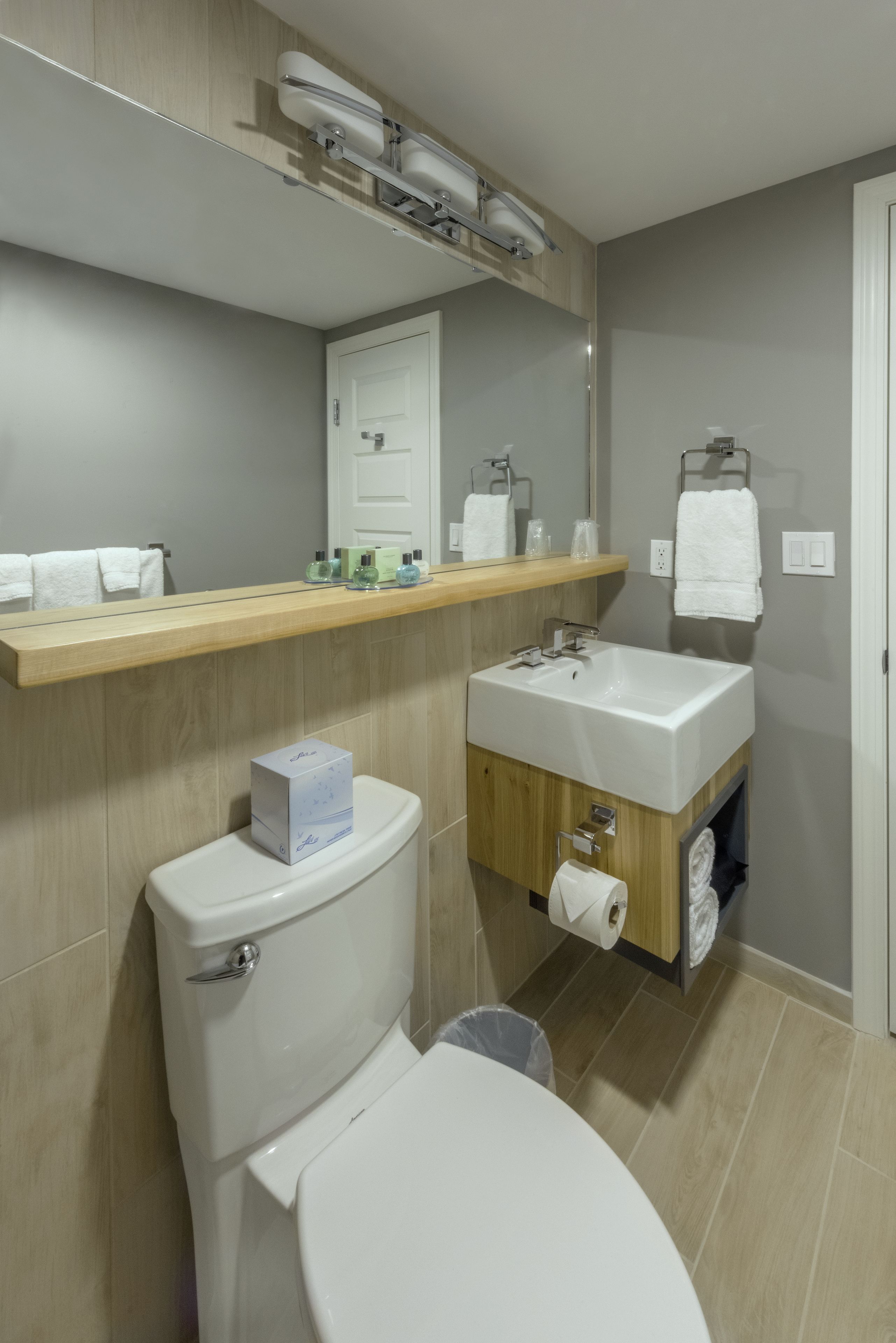A modern bathroom with a toilet, sink, and mirror. There is a shelf above the sink with toiletries.