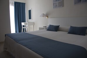 A neatly made bed with a blue bedspread and pillows, a white bedside table with a lamp, and a chair in a room with white walls and a window with blue curtains. - Sicania (Cullera)