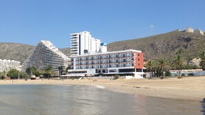 Un hôtel à étages, une plage en premier plan et une chaîne de montagnes en arrière-plan.
