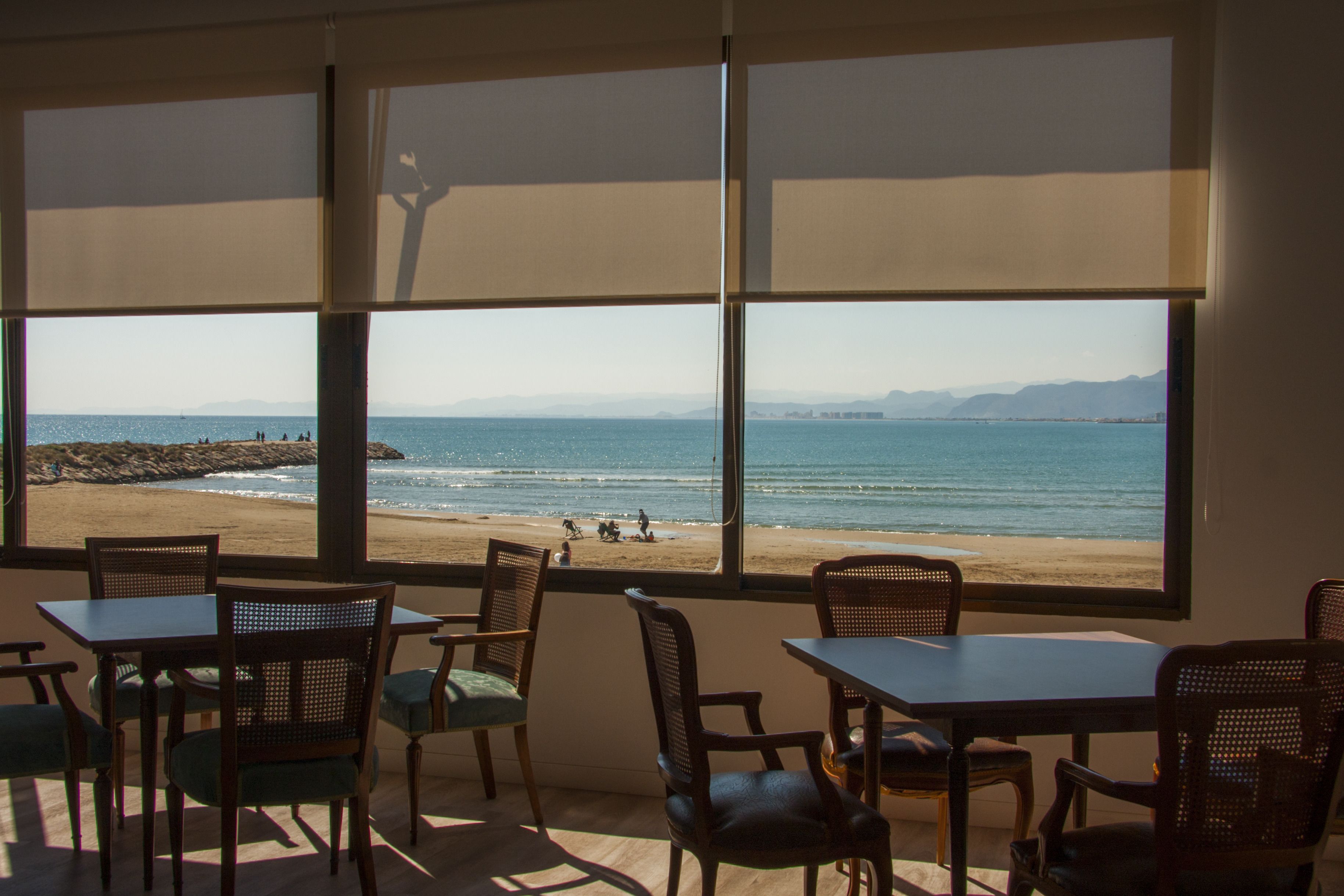 Una habitación con amplias ventanas que dan a una playa, amueblada con mesas y sillas de madera.