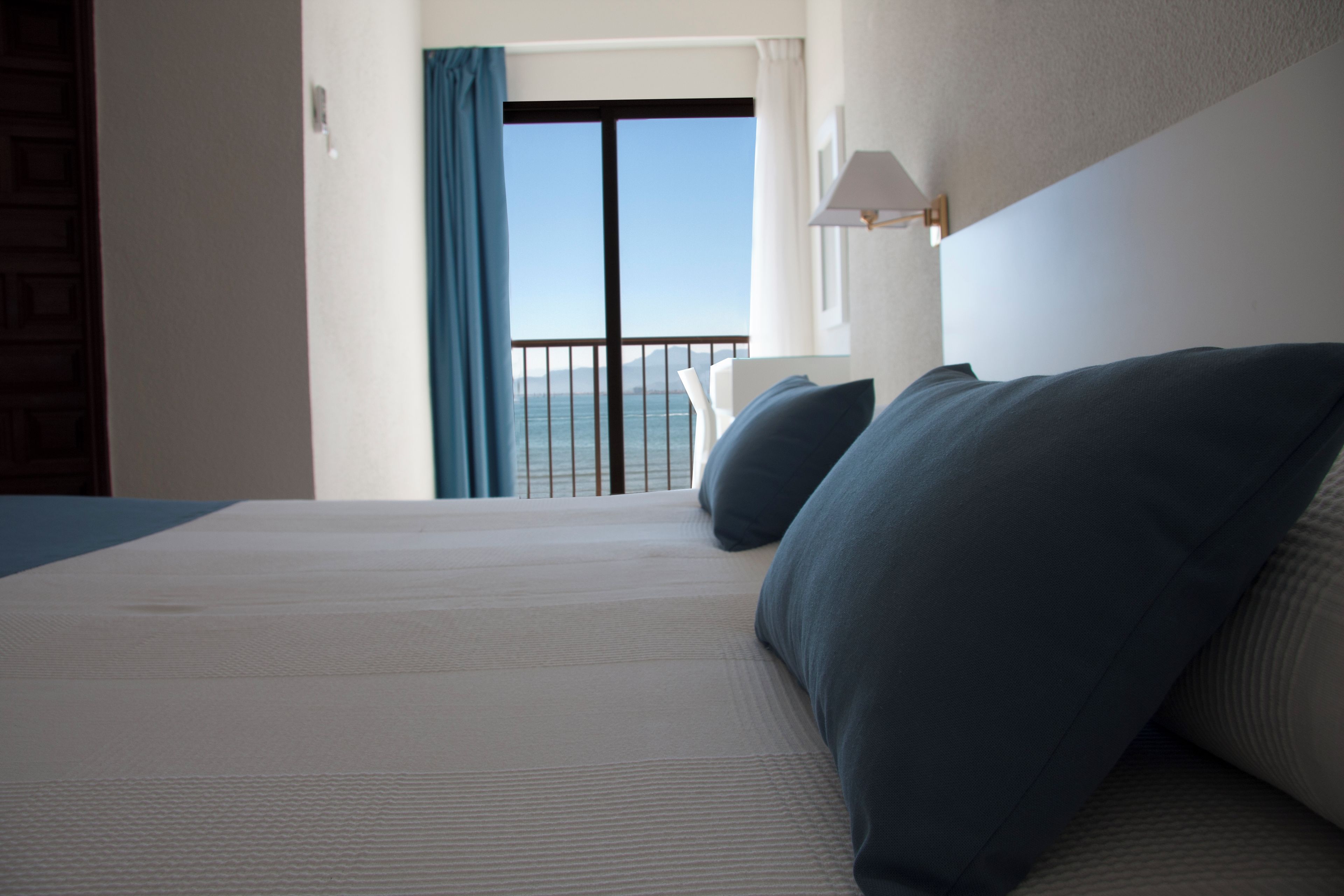 Een hotelkamer met een groot bed, uitzicht op de zee vanaf een balkon en een lamp aan de muur.
