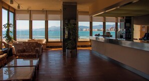 A hotel lobby with a large window overlooking the sea, a reception desk, and seating area. - Sicania (Cullera)