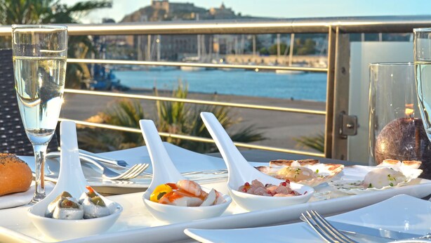 A table set for a meal with seafood dishes, a glass of water, and a view of a beach and water body in the background.