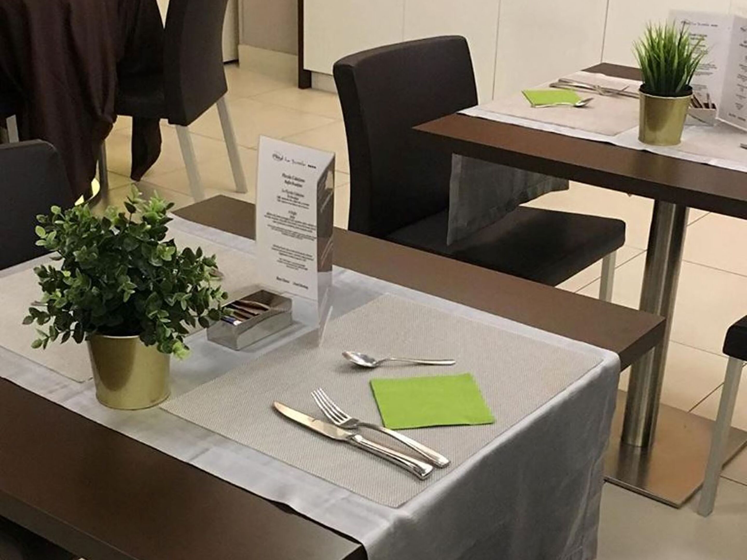 A hotel dining area with a table set for two, a menu stand, and a potted plant.