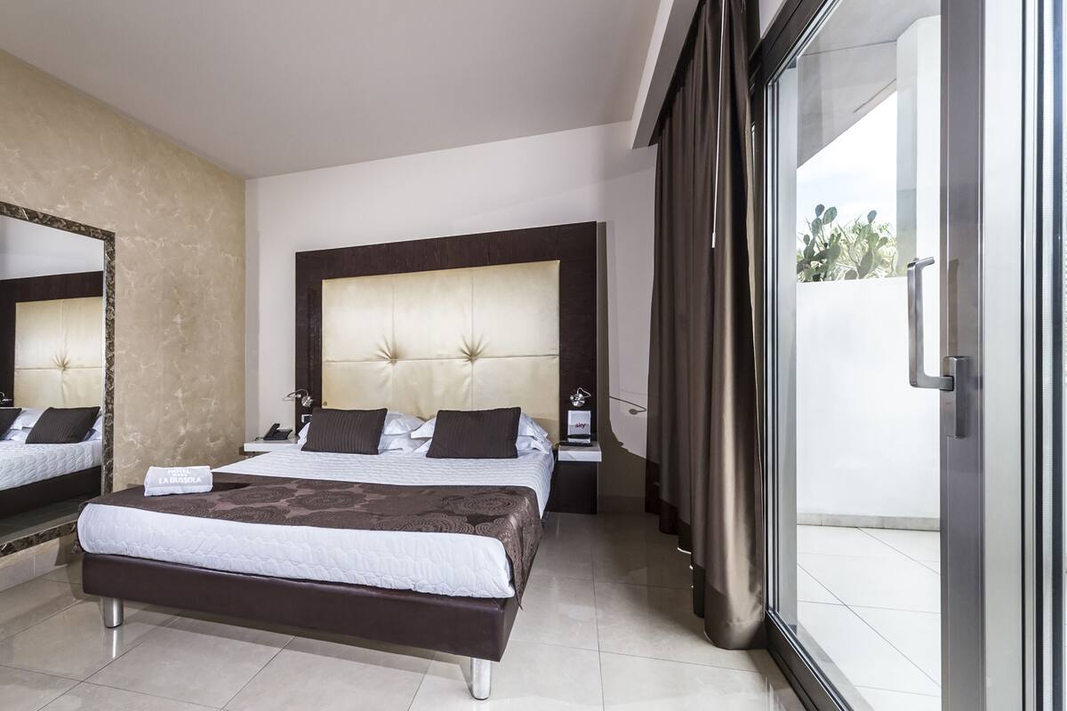 Quarto de hotel moderno com uma cama grande, mesa de cabeceira, espelho e vista através de uma porta de vidro.