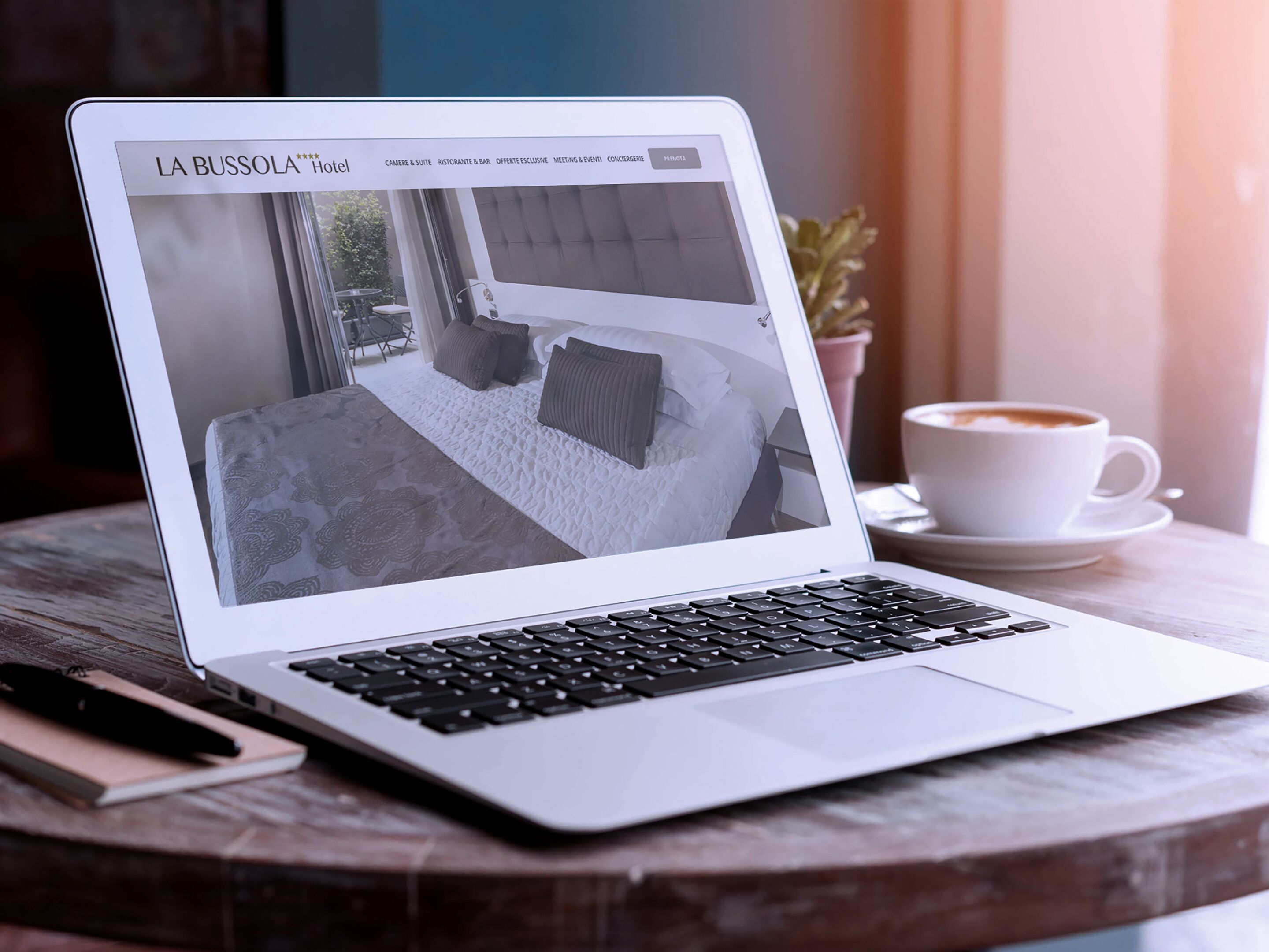 En bærbar computer viser et hotelwebsite med et moderne interiør, en kaffekop og en notesbog på et træbord.