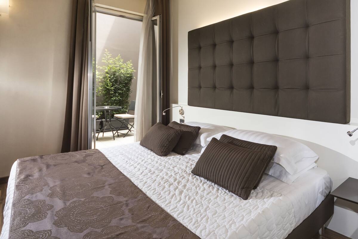 Quarto de hotel com uma cama grande, cabeceira, mesa de cabeceira e vista para um pátio externo.
