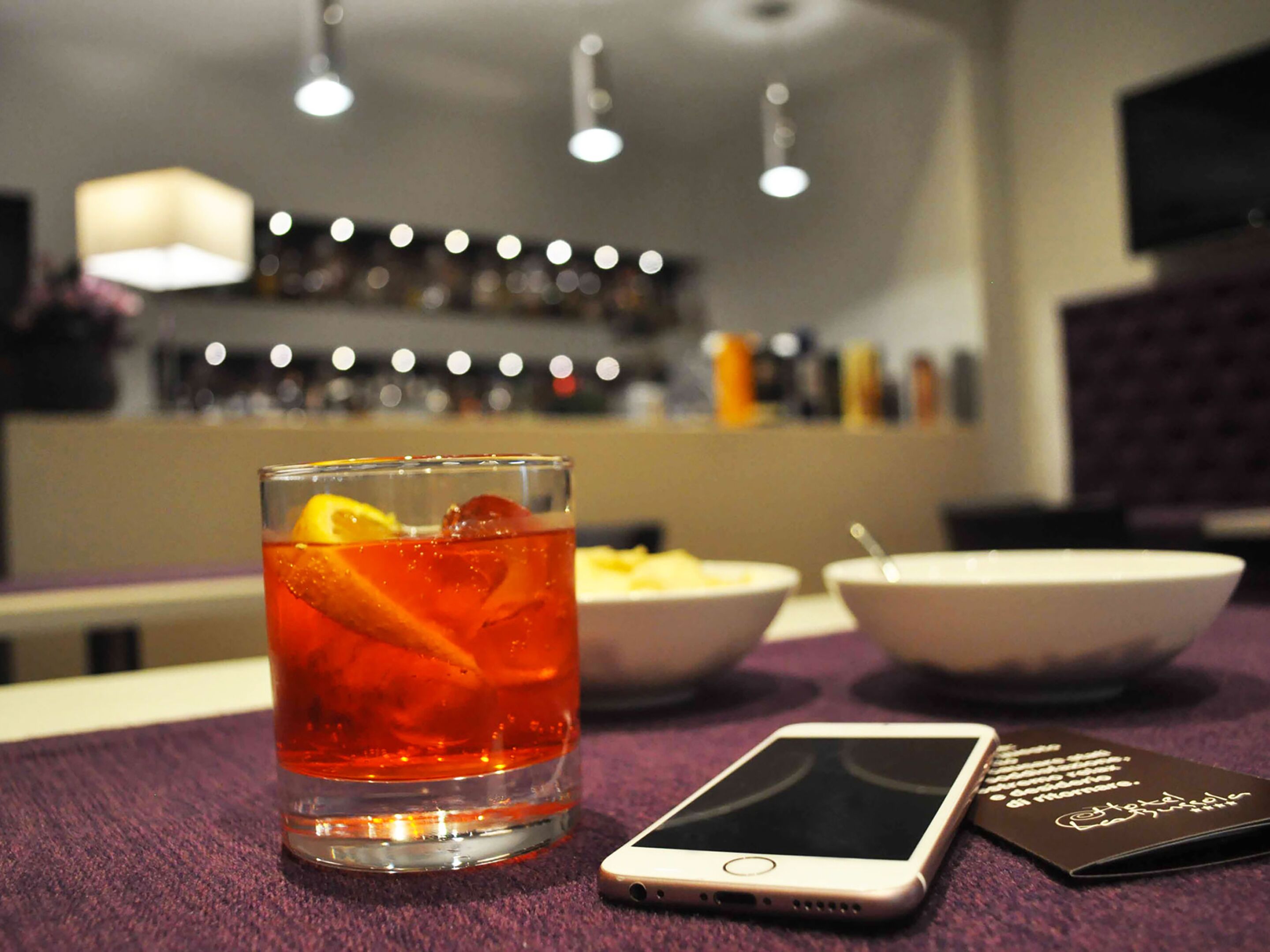 A glass of red beverage with an orange slice on a table, a smartphone, and two bowls of food in the background.