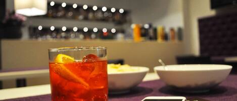 A glass of red beverage with an orange slice on a table, a smartphone, and two bowls of food in the background.