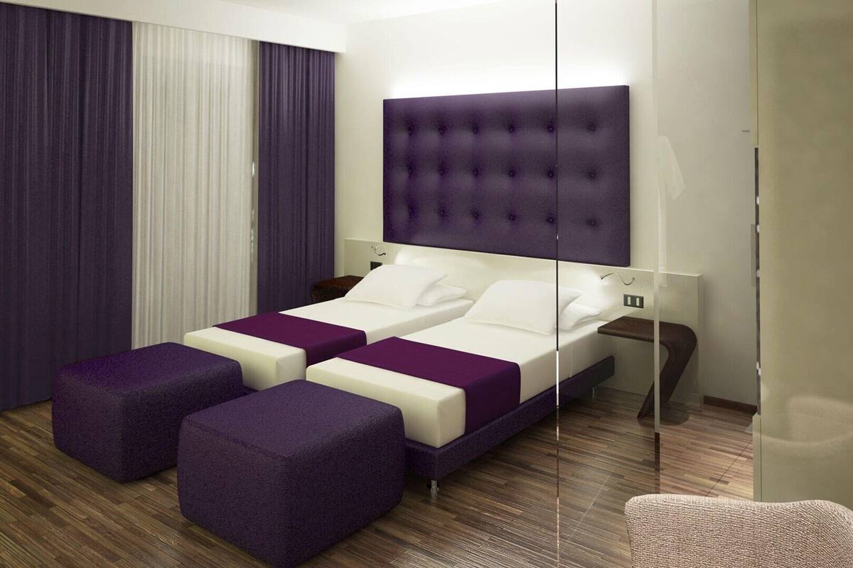 Quarto de hotel moderno com uma cama grande, duas mesas de cabeceira e um espaço de estar com pufes roxos.