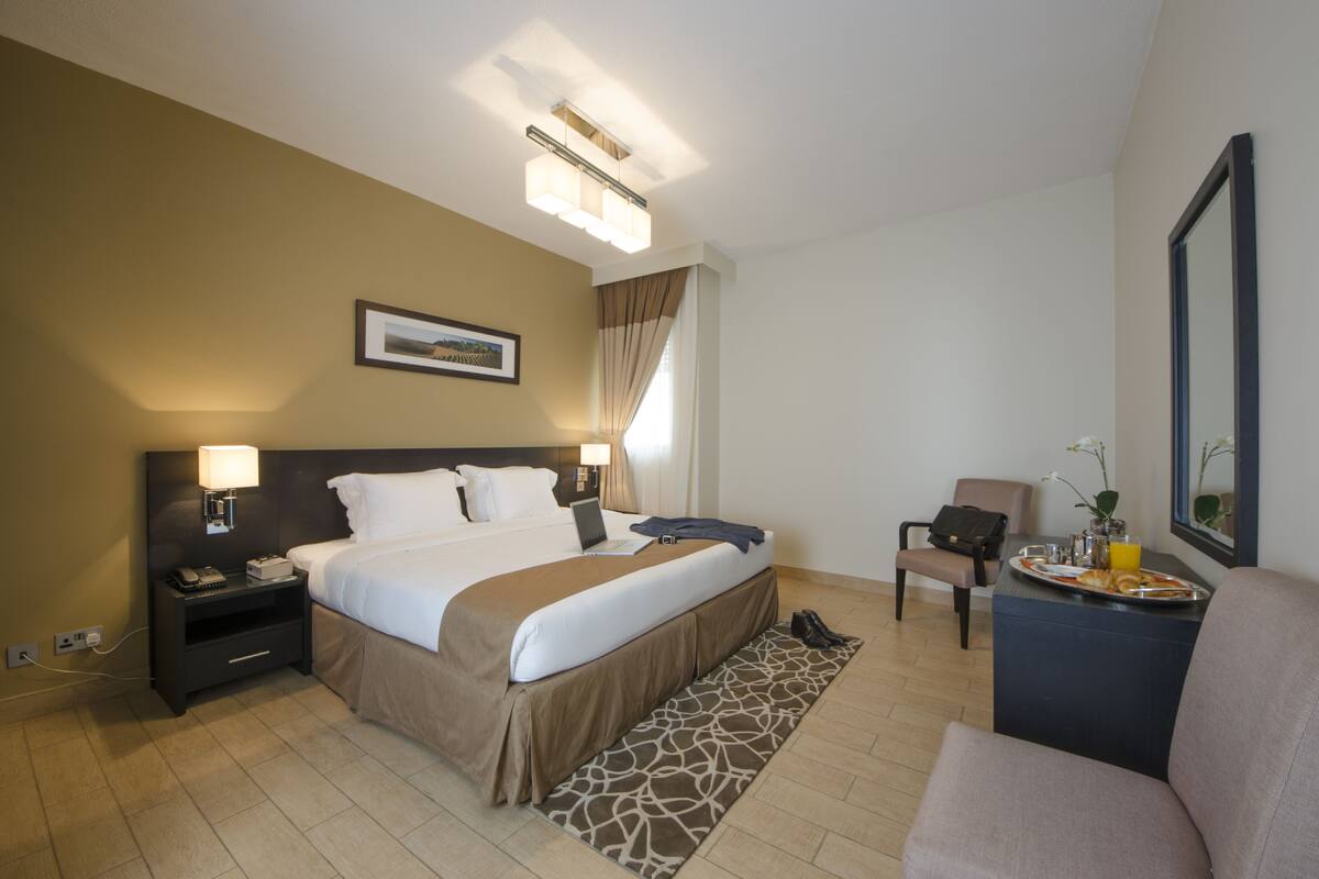 A hotel room with a bed, bedside table, lamp, chair, and a small table with a tray.