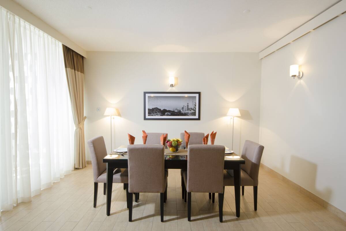 A dining area with a table set for four, surrounded by chairs, with a framed picture on the wall and two wall-mounted lamps.