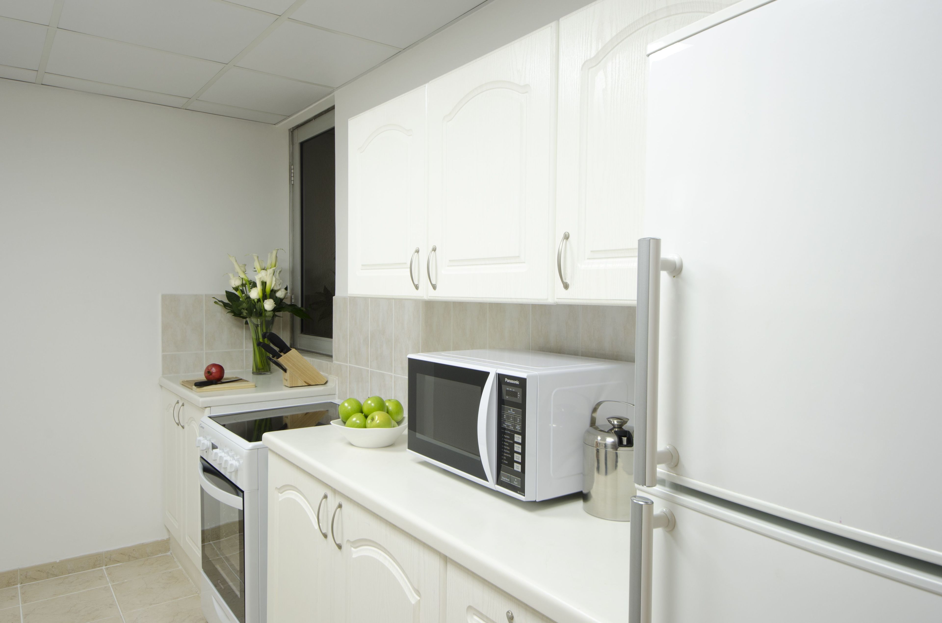 A kitchen with white cabinets, a microwave, a kettle, and a bowl of green apples.