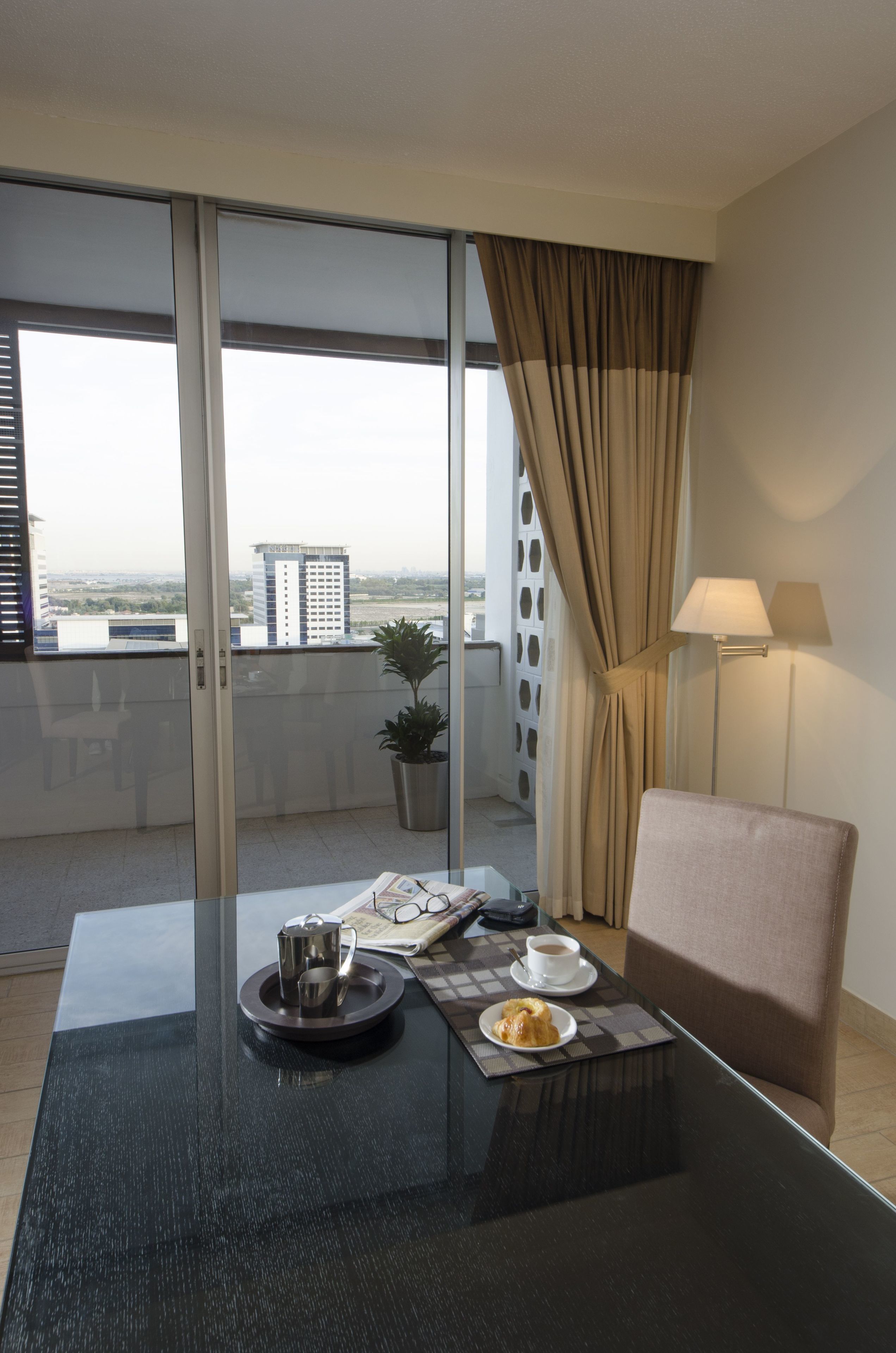 A modern hotel room with a balcony, a dining table with a coffee pot, a cup, and a plate of food, and a lamp on the side.