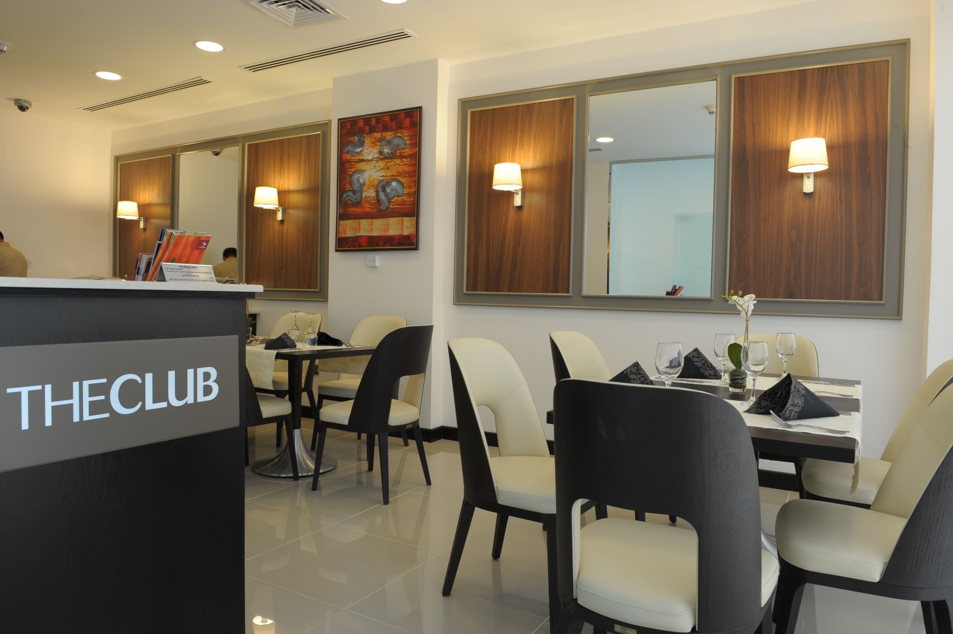A hotel lobby with a reception desk labeled "THECLUB", a dining area with tables and chairs, and a large mirror reflecting the room.