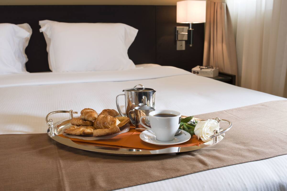 A neatly made bed with white linens, a tray with croissants, a cup of coffee, and a small bouquet of white flowers on a hotel room.