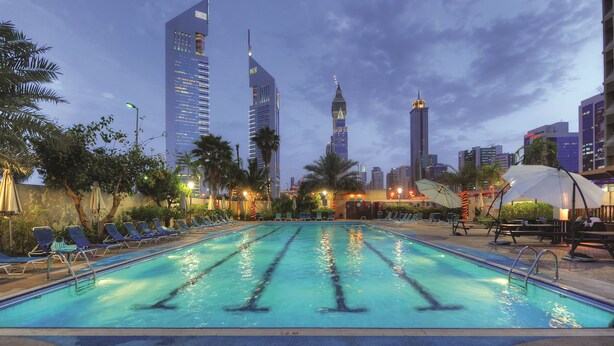 A swimming pool with lounge chairs and umbrellas, surrounded by palm trees and modern buildings in the background. - The Apartments Dubai World Trade Centre (Dubai)