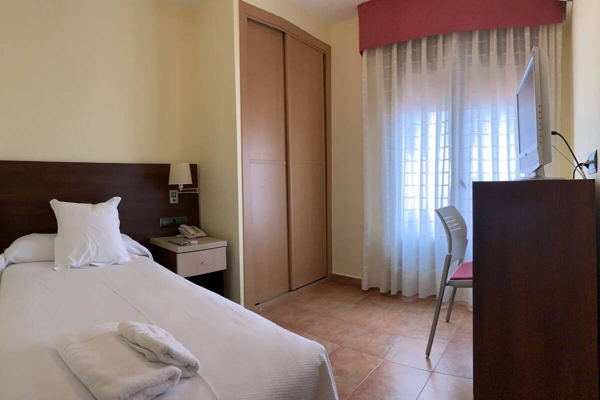 A hotel room with a bed, a desk with a telephone, a chair, a wardrobe, and a window with curtains.