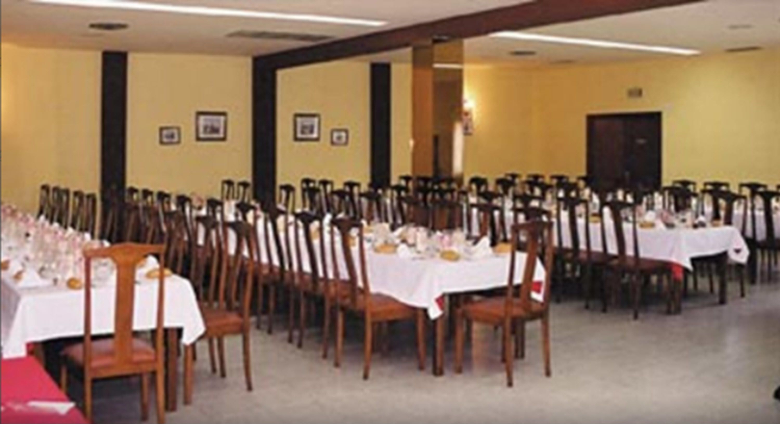 Een balzaal met rijen tafels gedekt voor een diner, houten stoelen en een gele wand met ingelijste foto's.
