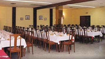 Een balzaal met rijen tafels gedekt voor een diner, houten stoelen en een gele wand met ingelijste foto's.
