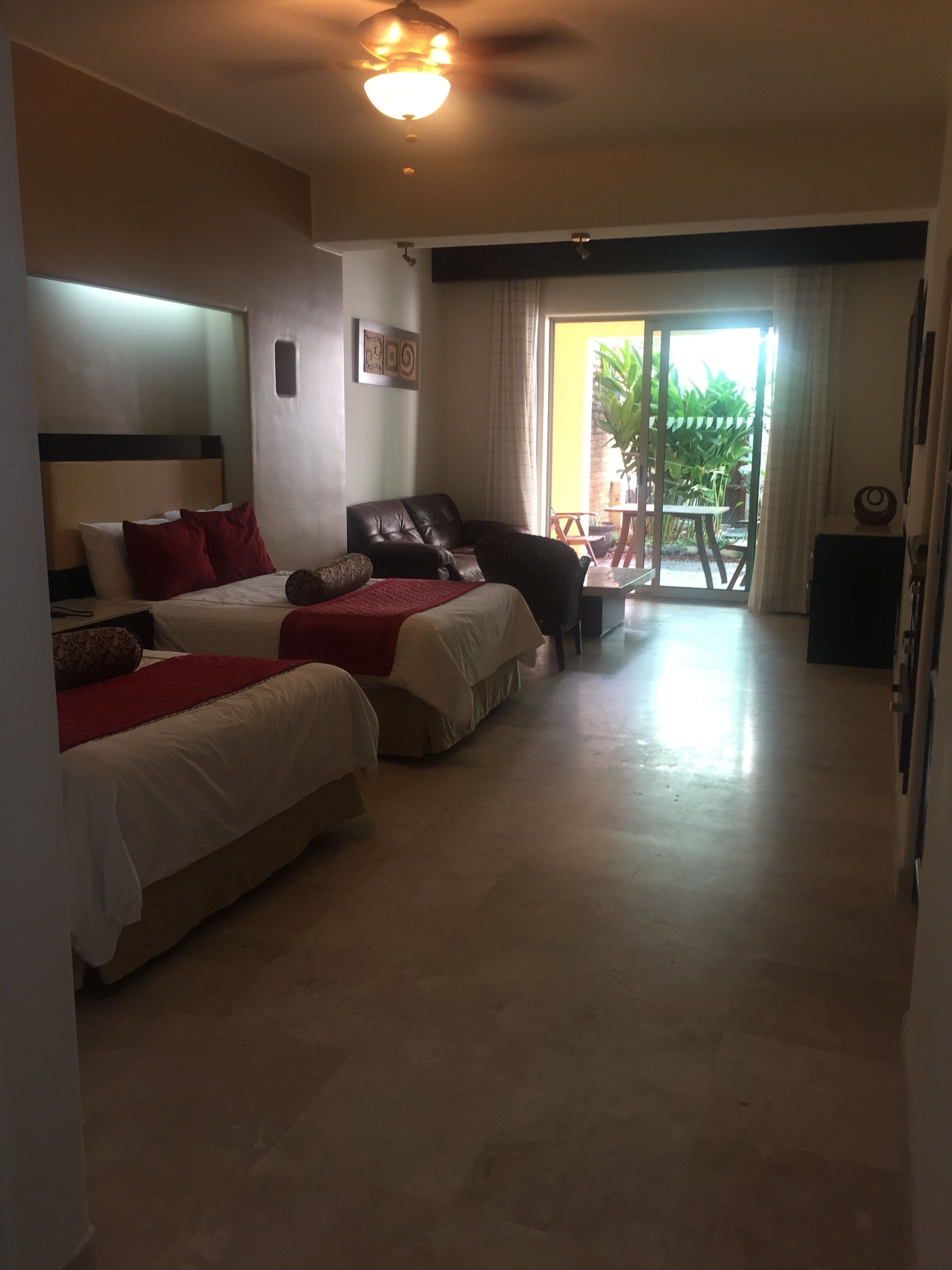 Habitación de hotel con dos camas, un sofá, un comedor con mesa y sillas, y un ventanal corredero que da a un espacio exterior.