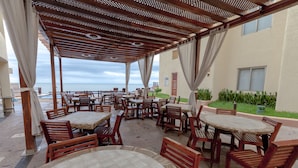Un patio al aire libre con varias mesas y sillas de madera, una estructura de pérgola y vista al océano.