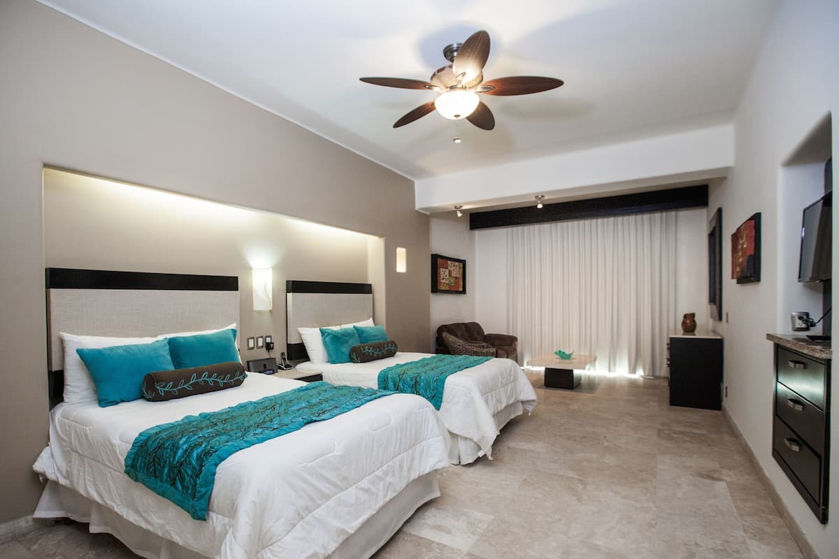 Habitación de hotel con dos camas, ventilador de techo, escritorio, silla y televisor.