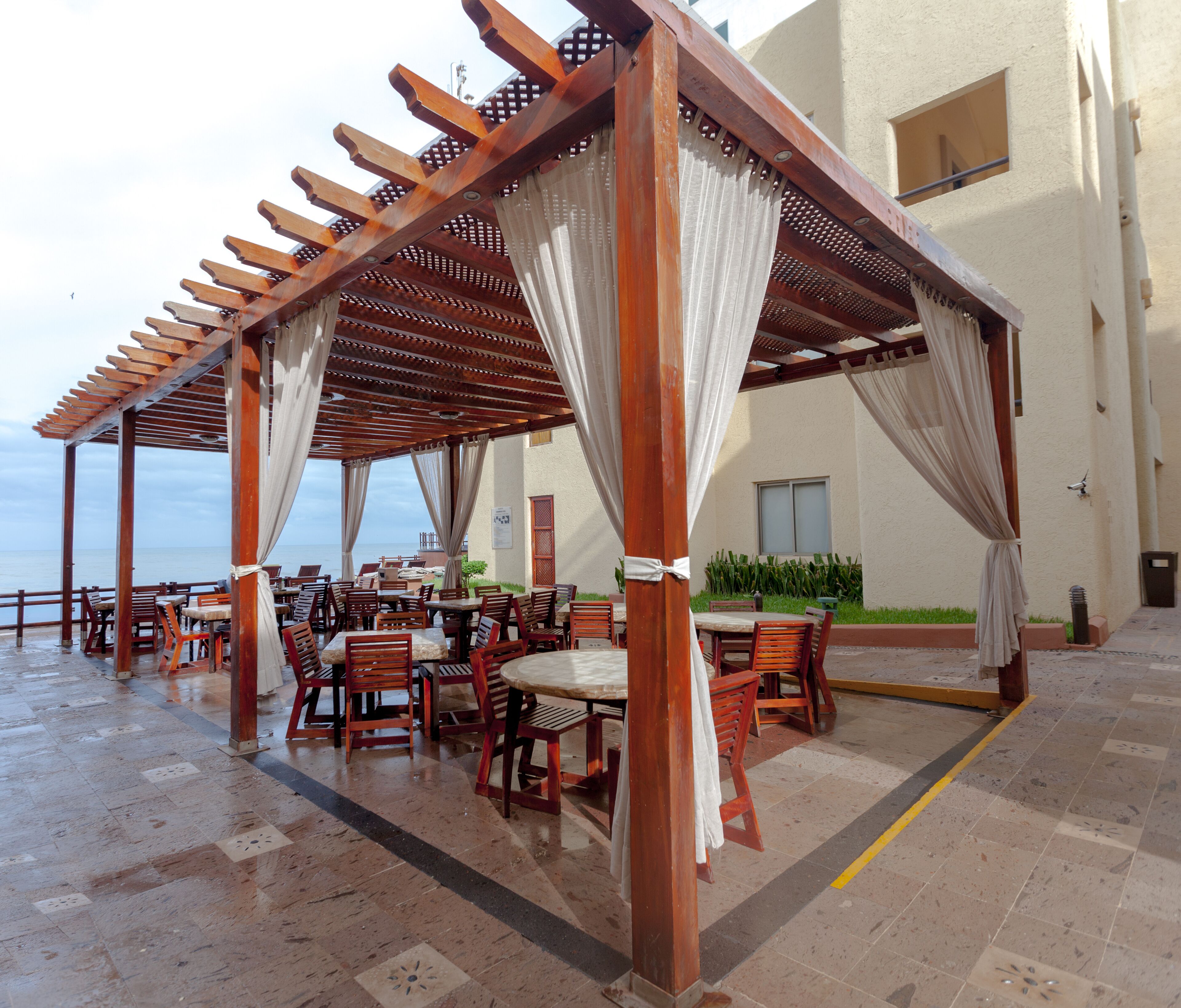 An outdoor patio with wooden tables and chairs, covered by a wooden pergola with white curtains.