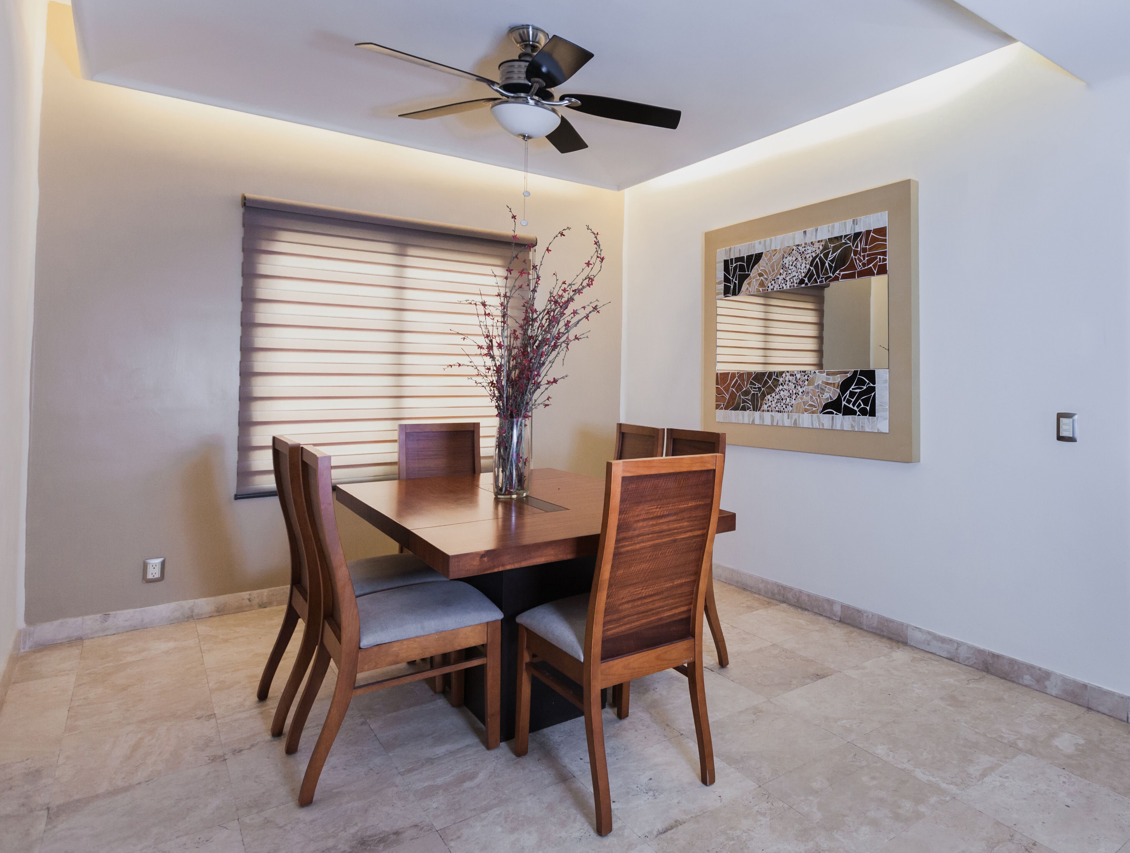 Un coin repas comprenant une table en bois, quatre chaises, un vase avec des branches, un ventilateur de plafond et un miroir fixé au mur.