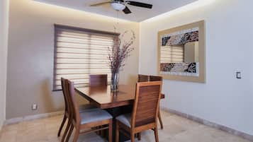 Un coin repas comprenant une table en bois, quatre chaises, un vase avec des branches, un ventilateur de plafond et un miroir fixé au mur.