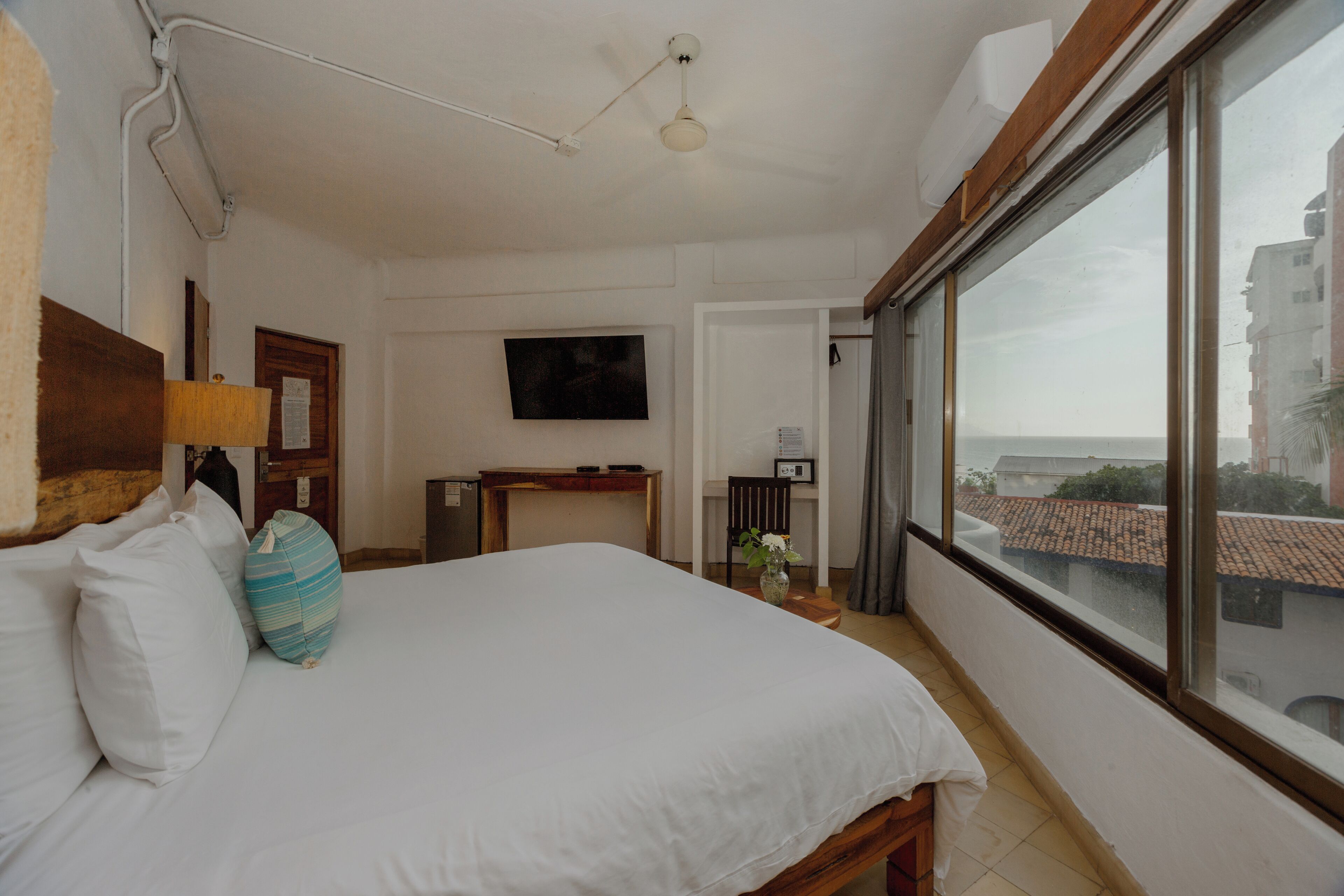A hotel room with a large bed, a TV mounted on the wall, a wooden headboard, and a view of the city through a large window.