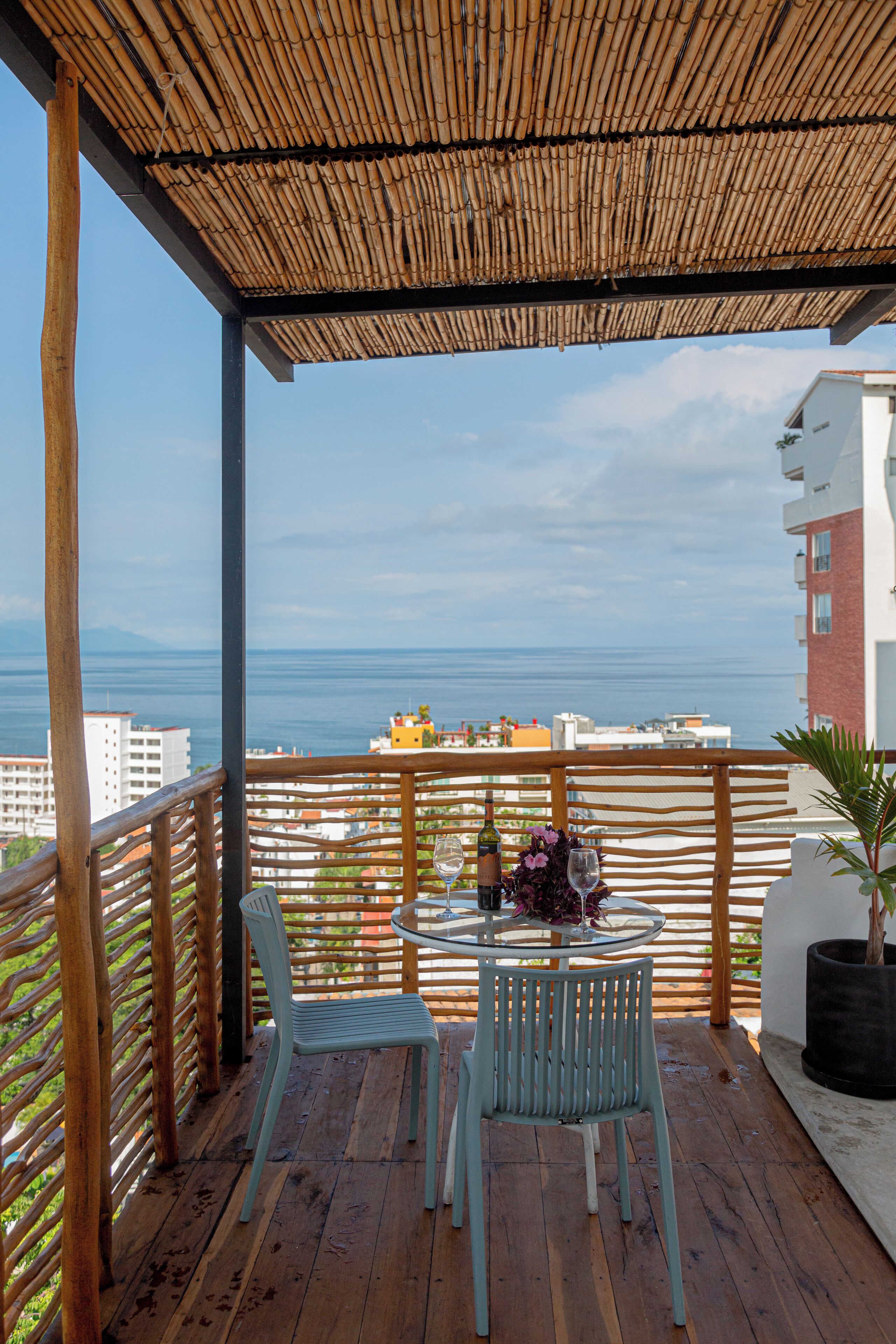 A wooden deck with a glass table and chairs, overlooking a city and the sea.