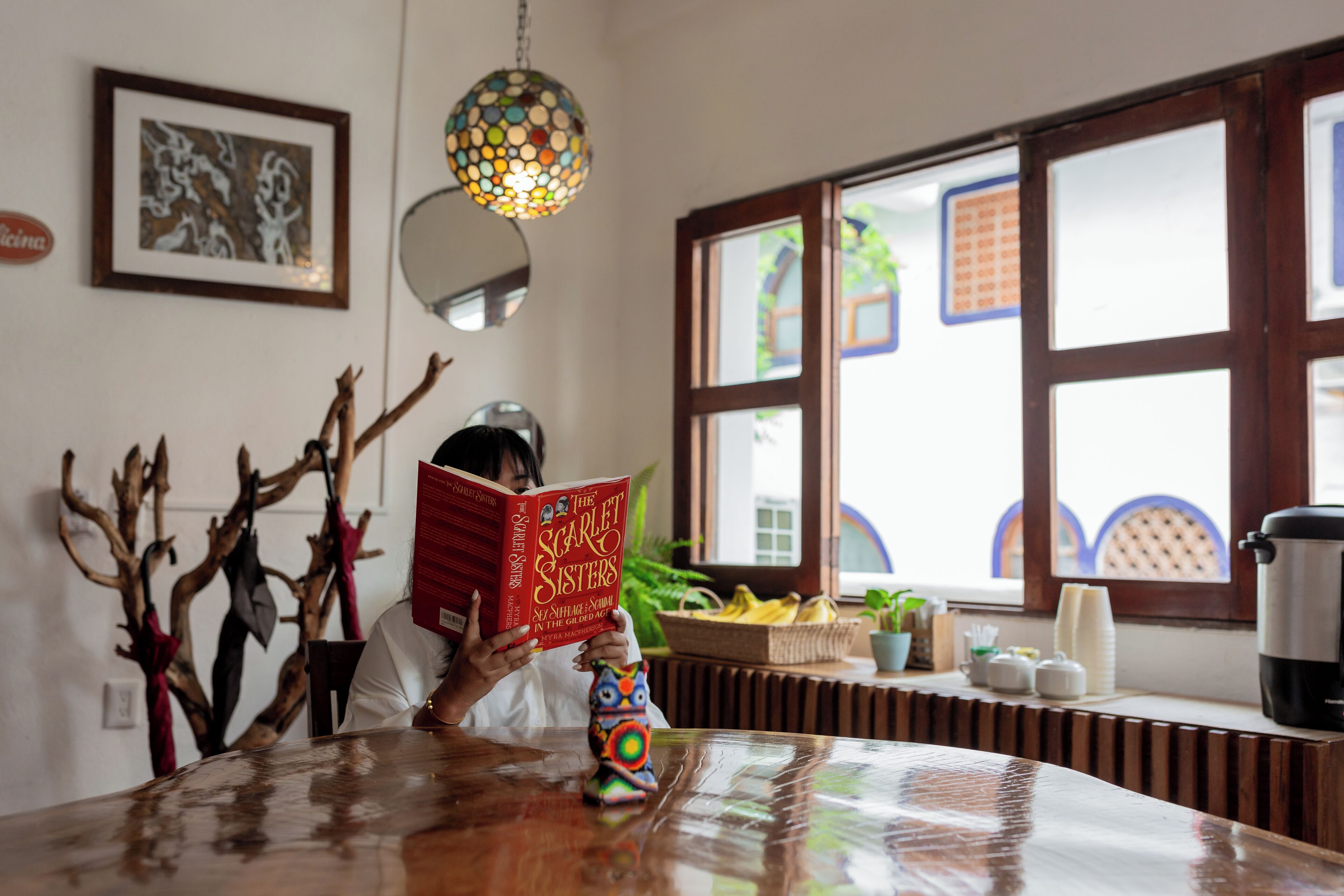 A person reading a book at a wooden table in a room with a large window, a decorative hanging light, and a framed picture on the wall.