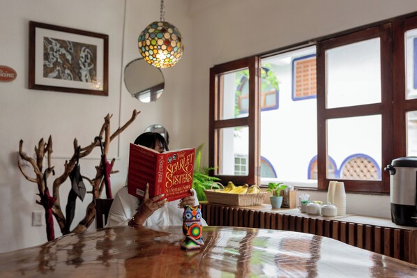 Una persona leyendo un libro en una mesa de madera en una habitación con un gran ventanal, una lámpara colgante decorativa y un cuadro en la pared.