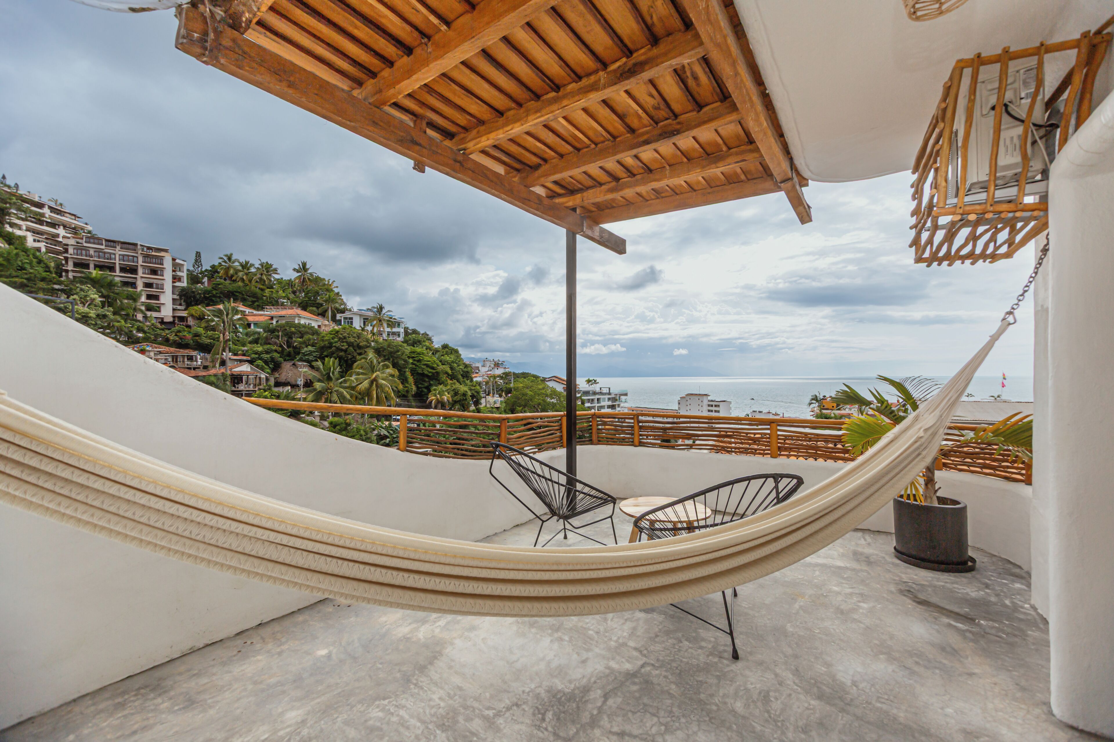 A hammock on a balcony with chairs, overlooking a beach and buildings.