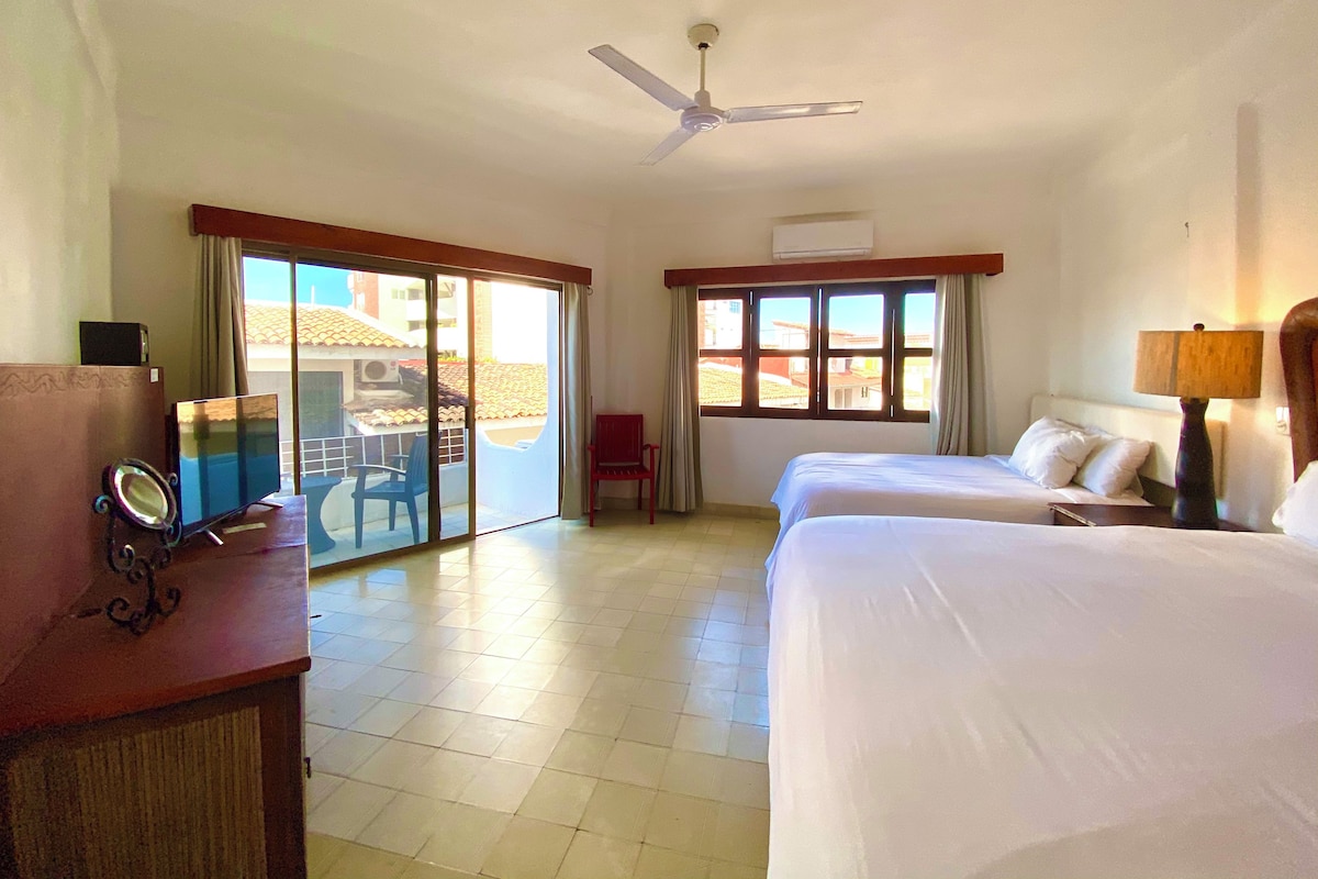 A hotel room with two beds, a ceiling fan, a desk with a computer, and a view of the outside through large windows.