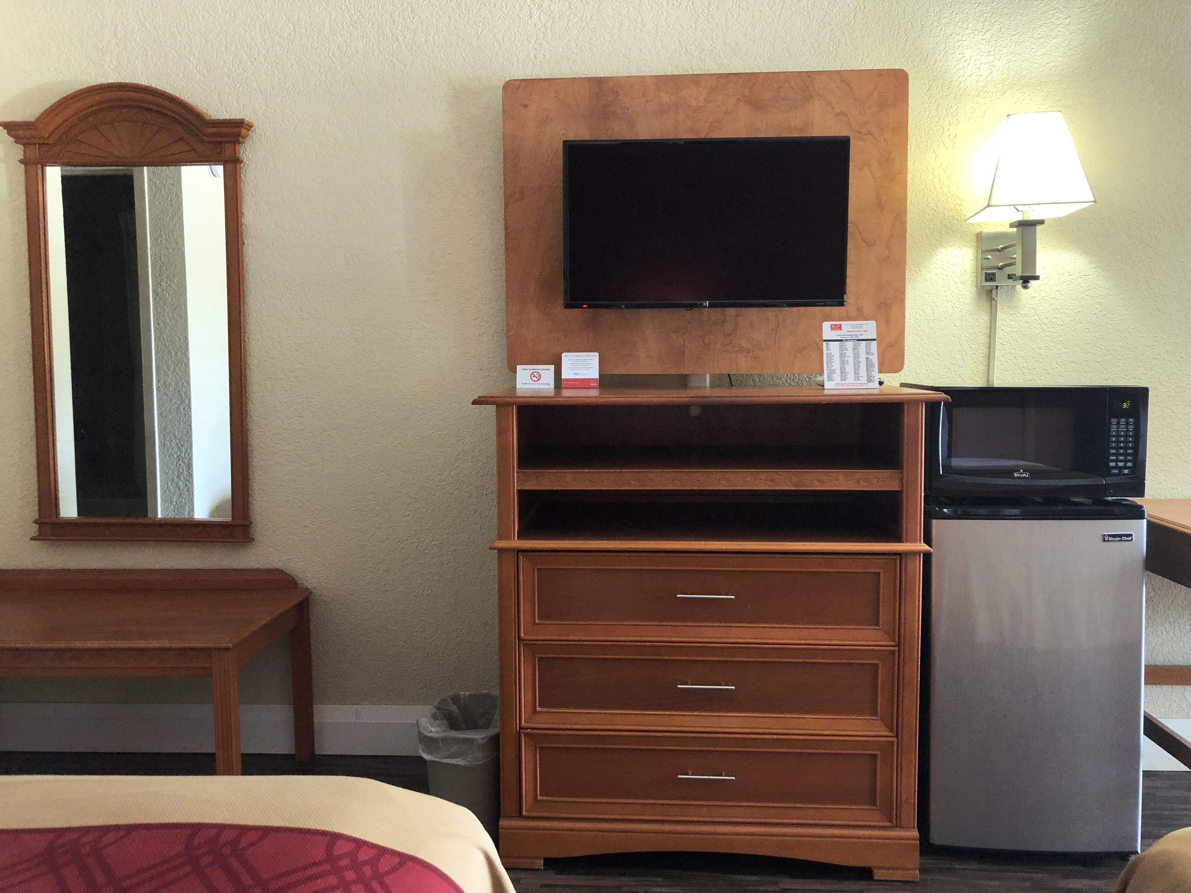 Habitación de hotel con cómoda de madera, televisor montado encima, microondas sobre un refrigerador, cesto de basura y cama con manta roja.