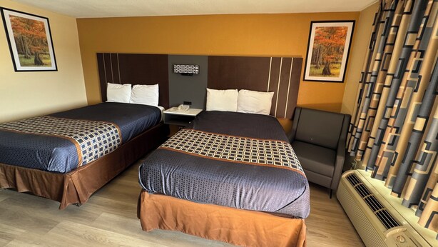 A hotel room with two beds, a desk, a chair, a heater, and two framed pictures on the walls. - Villa West Inn (Odessa)