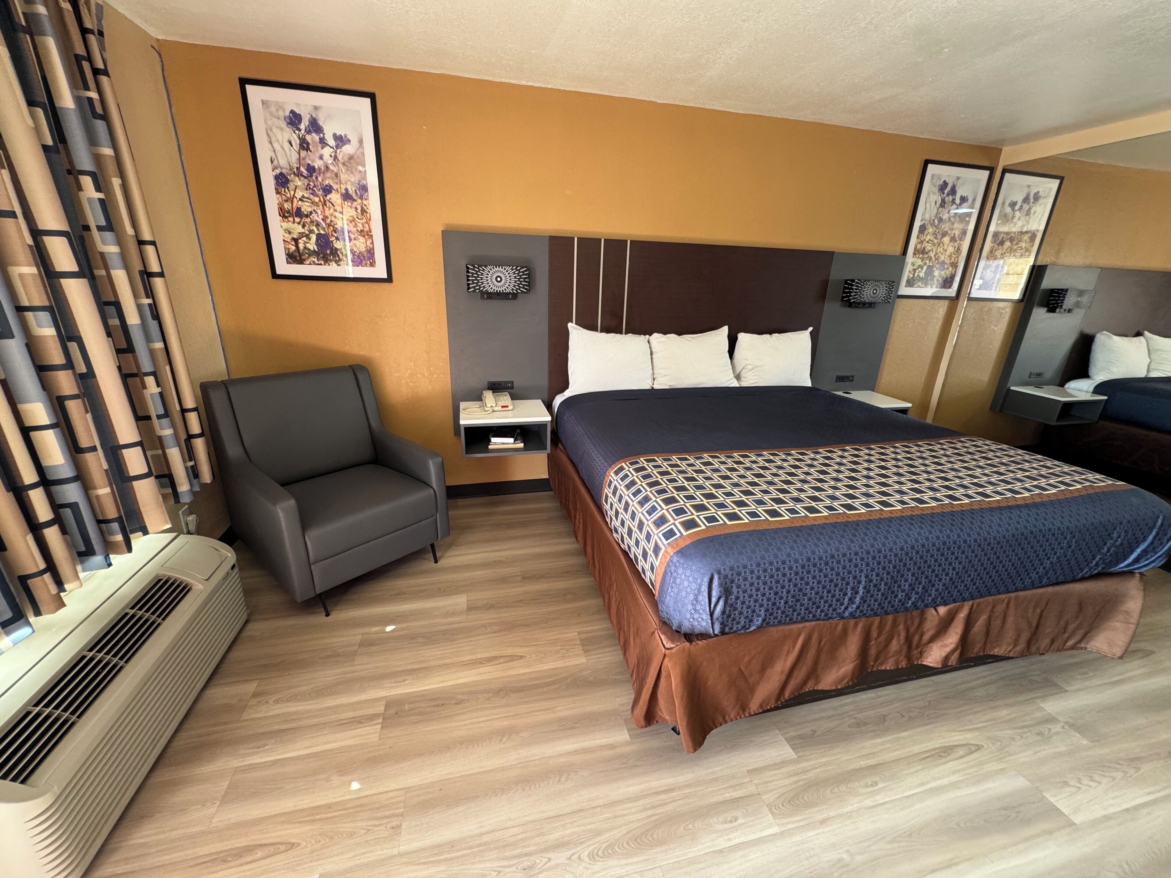 Una habitación de hotel con cama, una silla, una mesita de noche, un calentador fijado a la pared y cuadros enmarcados.