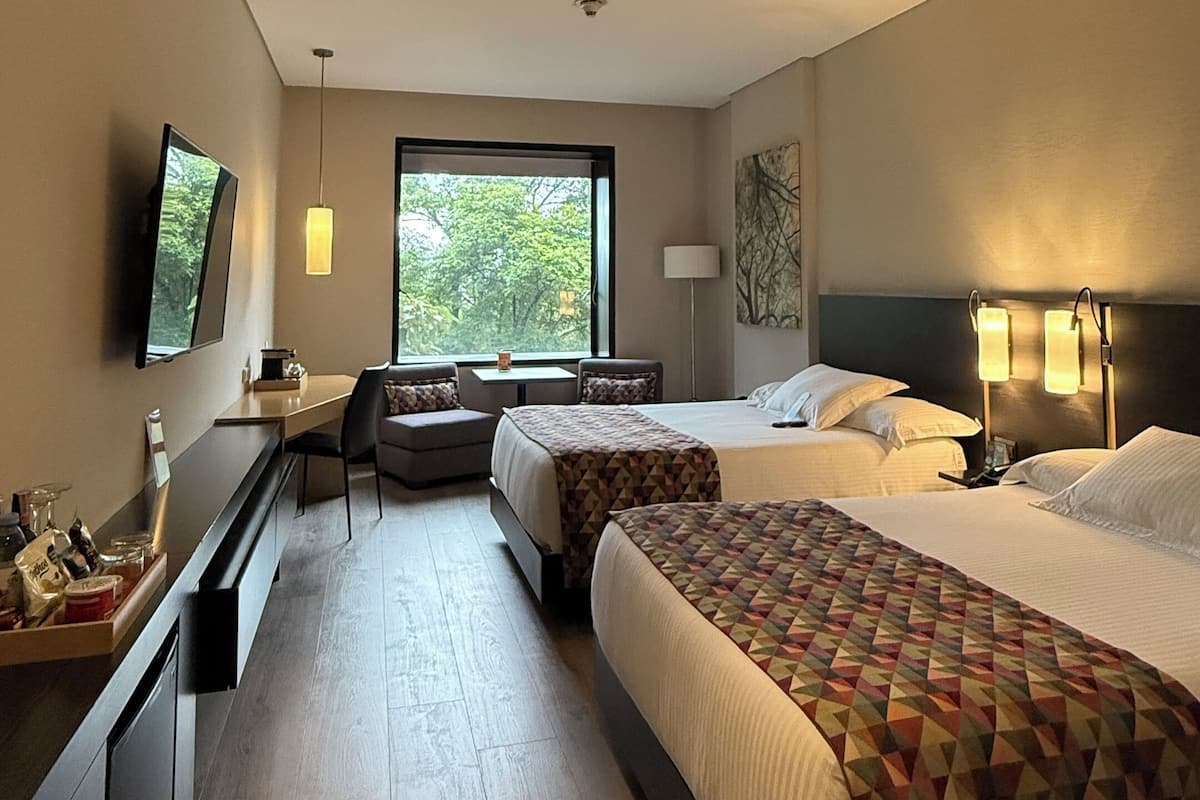 A hotel room with two beds, a desk, a chair, a TV, and a window with a view of trees.