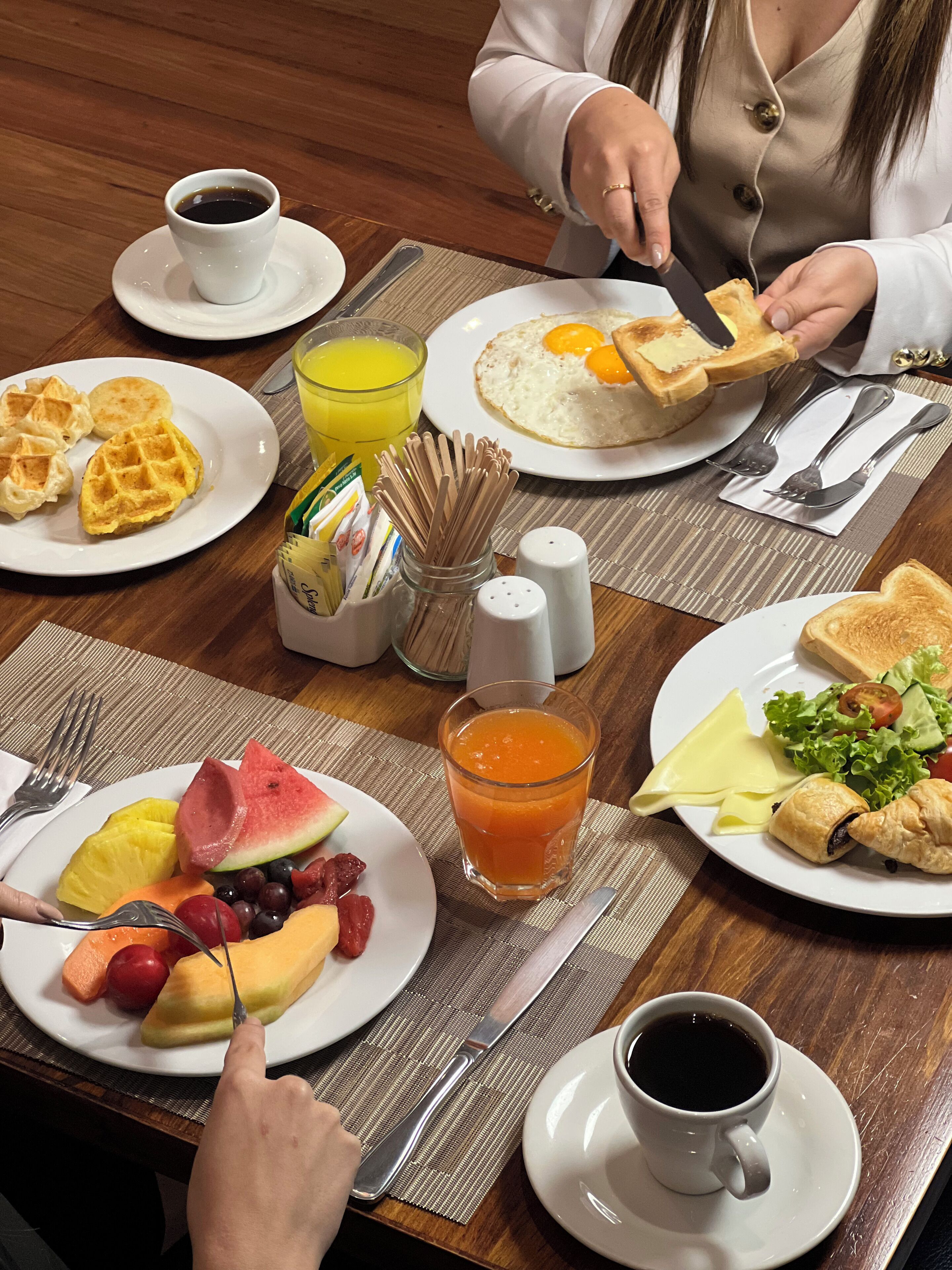 Una colazione con waffle, uova, pane tostato, frutta e succo in piatti, e una tazza di caffè.